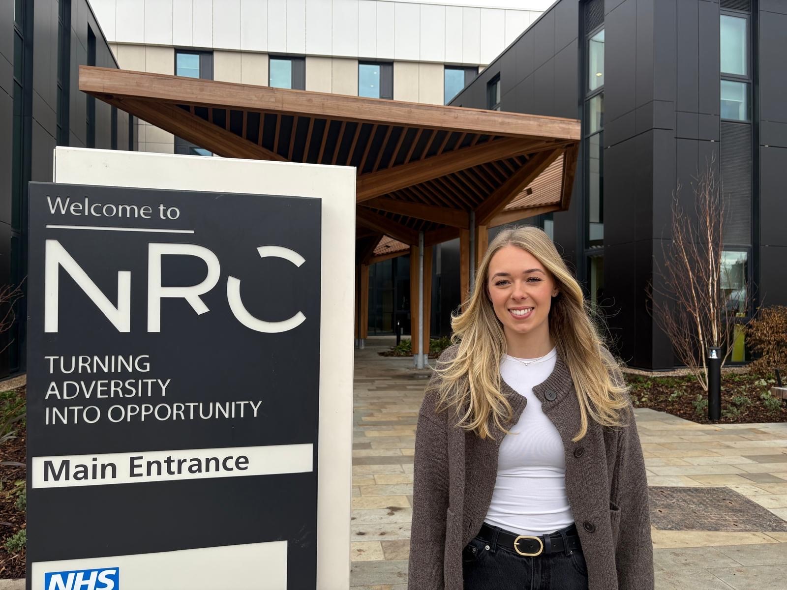 A woman with long blonde hair and stood in front of the NRC sign smiles at the camera